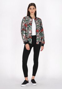 Bomberjack met bloemmotief in groen, rood en wit. Zwarte leggings en zwarte sneakers met witte zolen. Glad textuur van de stof.