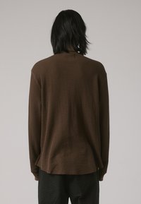 Chemise en maille gaufrée marron à manches longues avec une coupe décontractée, présentant un col rond et un détail de pli au dos, associée à un pantalon à motifs sombres.