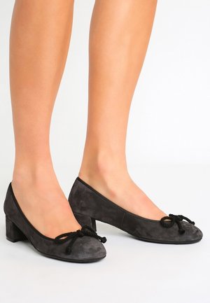 Pieds portant des chaussures noires en daim à talons bas carrés, bouts ronds et nœud tressé décoratif sur le dessus, debout sur une surface blanche.