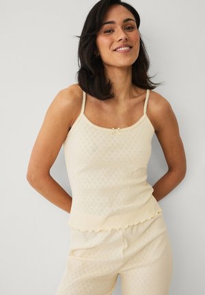 Kvinde med sort hår smilende, iført en cremefarvet ærmeløs top og matchende bukser med subtile hjerte mønstre, stående mod en ensfarvet baggrund.