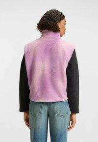 Fleece-vesti, hvor gradienten er i rosa og lavendel, høy krage, ermløs design, myk tekstur, lagd over en svart langermet skjorte, sammen med blå jeans.