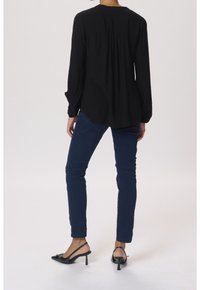 Personne portant un blouse noire à manches longues, un jean slim bleu foncé et des escarpins à talons noires, se tenant devant un fond clair.