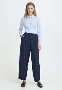 Hele sinine nööpidega pluus kraega, kombineeritud mereväe laia säärega pükstega. Mõlemal riideesemel on sile tekstuur ja pluusil on pikad varrukad.