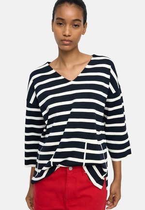 Kobieta ubrana w czarno-białą, poziomo paskowaną długorękawową bluzkę z dekoltem w serek i czerwone spodnie, stojąca na jednolitym tle.