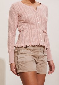 Kobieta ubrana w różowy, długi sweter na guziki z wiązanym w pasie paskiem oraz beżowe szorty z mankietami na jednolitym tle.