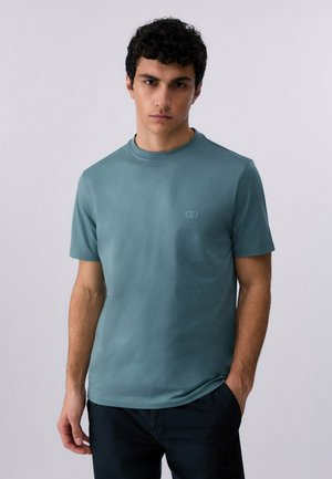 Giovane uomo con capelli corti e ricci, che indossa una maglietta girocollo tinta unita color teal e pantaloni neri, in piedi contro uno sfondo chiaro e uniforme.