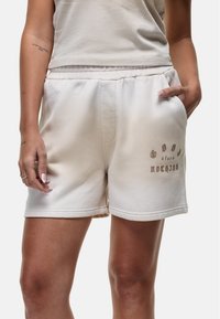 Pantaloni corti in misto cotone beige con vita elasticizzata, tasche laterali e design testuale stampato in marrone sul lato sinistro inferiore.