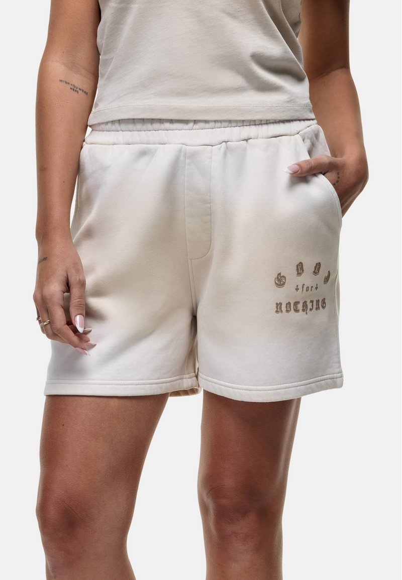 Pantaloni corti in misto cotone beige con vita elasticizzata, tasche laterali e design testuale stampato in marrone sul lato sinistro inferiore.