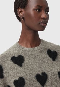 Maglione lavorato a maglia di colore grigio chiaro con evidenti motivi a cuore neri. Collo rotondo, texture morbida e vestibilità rilassata. Design minimalista.