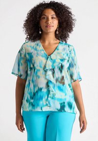 Blusa leggera con motivo blu e verde, colletto a V, frontale con volant e maniche corte, abbinata a pantaloni blu brillante.