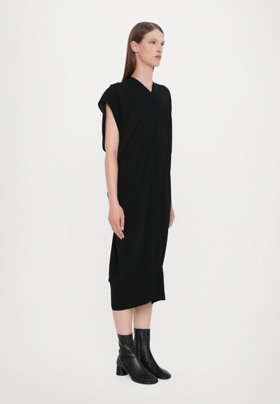 Black, knee-length dress with a V-neck and short sleeves, made from soft fabric. Model wearing black ankle boots. Simple, fluid design.