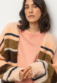 Strickjacke mit rosa, braunen und marineblauen Streifen über einem hellrosa T-Shirt, auf dem "SENSES. THE LABEL" in kleiner Schrift gedruckt ist. Weicher Stoff.