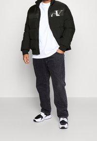 Sort dunjakke med lodret quiltet design, med hvidt logo på venstre bryst, båret over en hvid t-shirt og mørke jeans.