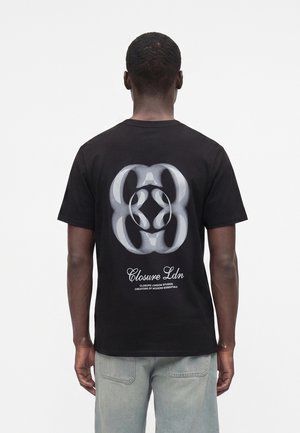 Uomo di spalle, indossa una T-shirt nera con una grande grafica bianca astratta e la scritta “Closure Ldn” sul retro, jeans in denim chiaro.