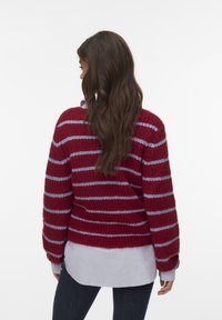 Eine Frau mit langen braunen Haaren trägt einen roten Strickpullover mit hellblauen horizontalen Streifen über einem hellgrauen Hemd und dunklen Jeans und zeigt mit dem Rücken.