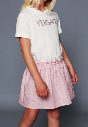 Niña con camiseta blanca con el logo de Versace y falda floral rosa pálido sobre un fondo gris, de pie con los brazos relajados a los lados.