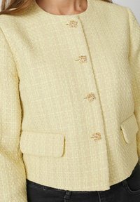 Svetlo rumena tweed jakna z okroglo ovratnikom, z zlatimi detajli, štirimi gumbi in dvema sprednjima žepoma. Ima teksturirano tkanino.