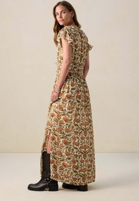 Bloemige maxi-jurk met ruches mouwen, groene basis en bedrukt met oranje en roze bloemen; heeft een hoge split tot aan de dij en wordt gedragen met zwarte laarzen.