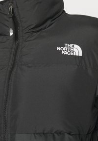 Czarna kurtka puchowa z wysokim kołnierzem, zapinana na zamek, z białym logo "The North Face" na lewej piersi. Gładki, ocieplany materiał.