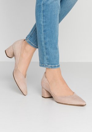 Kvinde iført beige ruskind blokhælssko kombineret med blå denimjeans, stående på en glat, lysegrå gulv.
