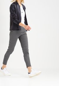 Svart lättviktsjacka över en vit skjorta, parat med åtsittande svarta och vita hundtandsmönstrade byxor och vita träningsskor.