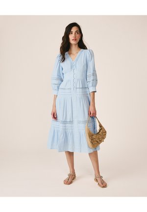 Femme portant une robe midi bleu clair avec des manches bouffantes, tenant un sac à main beige avec des détails floraux, et portant des sandales ornées.