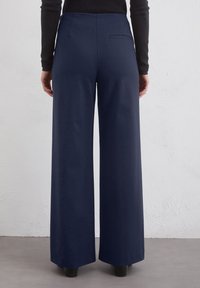 Pantaloni largi de culoare bleumarin, confecționați dintr-un material neted, cu talie înaltă și un singur buzunar la spate. Țesătura se termină deasupra gleznei.