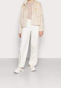 Veste en fausse fourrure crème avec un design légèrement court, deux poches avant, associée à un pantalon blanc à jambes larges et des baskets beige.