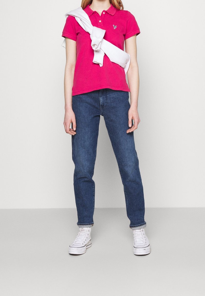 Rosa Polohemd mit kleinem Logo, dunkelblaue Jeans und weiße Sneakers. Ein weißer Pullover ist über die Schultern gelegt.