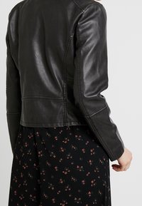 Giacca di pelle nera con un design aderente, caratterizzata da una texture liscia, cuciture sulle spalle e una lunghezza corta, abbinata a un vestito floreale.