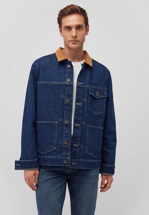 Jeansjacke mit einem braunen Cordkragen, großen Fronttaschen, Metallknöpfen und sichtbaren Nähten. Über einem weißen Hemd getragen.