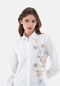 White button-up shirt featuring a floral watercolor design in soft colors on one side. Made of cotton with a classic collar and long sleeves.
