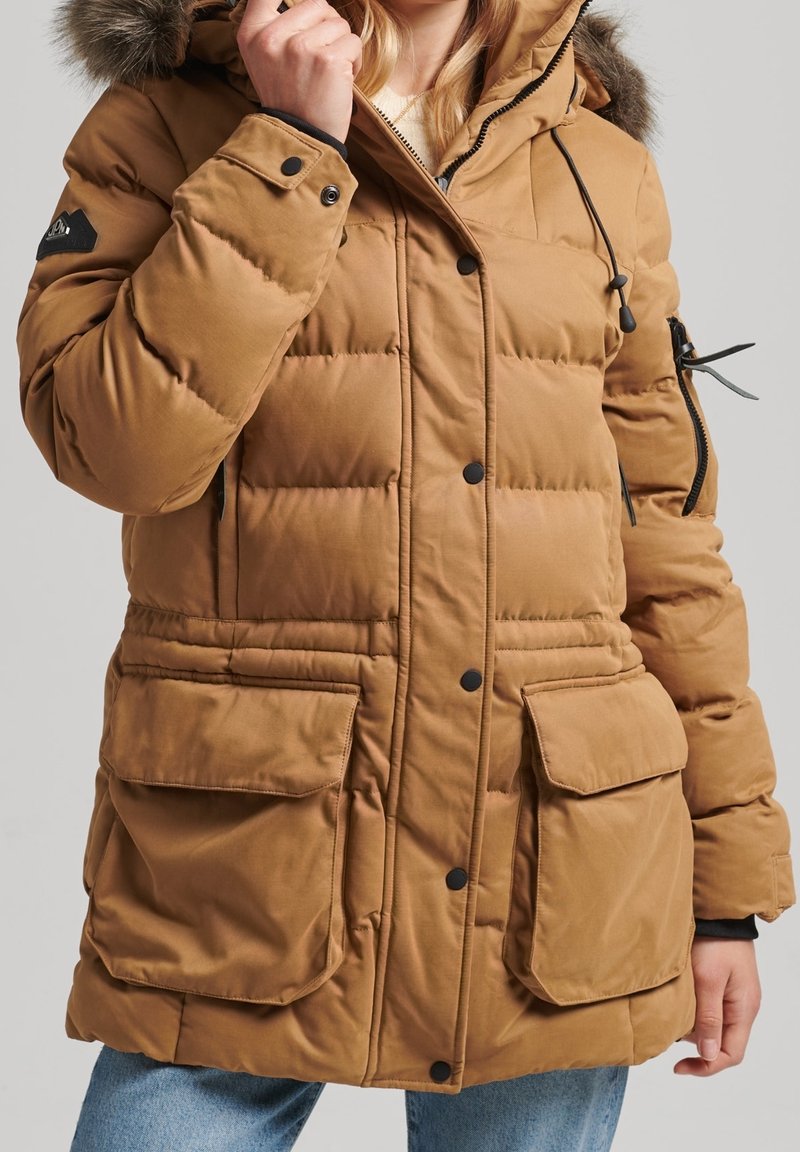 Veste parka matelassée marron avec capuche bordée de fourrure, grandes poches avant, fermeture à boutons sur le devant et cordons élastiques à la taille.