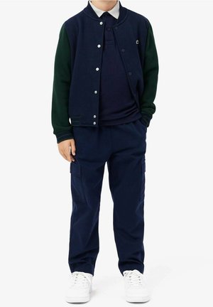 Chaqueta universitaria azul marino y verde con botones de presión, sobre una camiseta polo azul marino, combinada con pantalones cargo azul marino y zapatillas blancas.