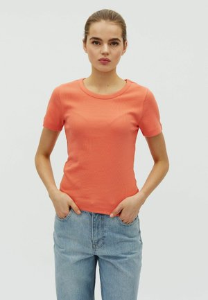 Noor nahaga noor naine, kes kannab korallivärvi lühikeste varrukatega särk ja helesiniseid pükse ning kellel on käed taskutes.