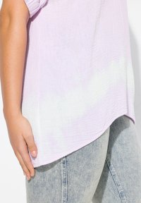 Top di un delicato viola chiaro, con una texture e un effetto sfumato sottile, abbinato a jeans attillati di un azzurro chiaro. Dettagli notevoli includono una linea dell'orlo curva e maniche arricciate.