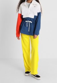 Windbreaker-Jacke in Weiß, Rot und Marineblau mit Kordelzug in der Taille; kombiniert mit leuchtend gelben Hosen und schwarzen Sneakers. Verfügt über ein Halb-Reißverschluss-Design.