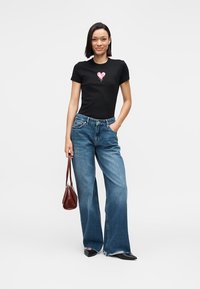 Camiseta negra de manga corta con un gráfico de corazón rosa, combinada con vaqueros de pierna ancha en azul y un pequeño bolso burdeos.