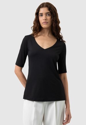 Femme aux longs cheveux bouclés portant un haut noir à col en V à manches mi-longues et un pantalon blanc, debout devant un fond uni.