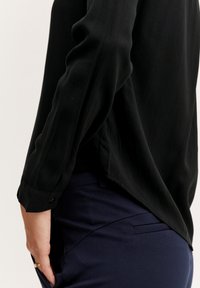 Blusa nera a maniche lunghe con texture liscia, dotata di un orlo curvo e polsini con bottoni, abbinata a pantaloni sartoriali blu navy.