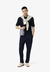 Maglione lavorato a maglia navy, sciarpa a righe crema, pantaloni navy e sneakers chiare. Borsa a tracolla grigia con logo impressa. Outfit casual.