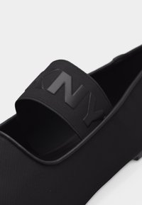 Μαύρο παπούτσι χωρίς κορδόνια με υφασμάτινη υφή και φαρδιά ελαστική λωρίδα στο πάνω μέρος, με ανάγλυφα στυλιζαρισμένα γράμματα.