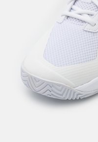 Białe buty sportowe z teksturowaną siateczką na cholewce, gładkim gumowym czubkiem, wzorem w jodełkę na podeszwie oraz płaskimi sznurowadłami.