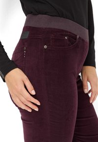 Burgunderfarbene Cordhose mit einem breiten, gerippten Bund, vorderen Taschen und dekorativen Metallakzenten an der Seite. Insgesamt glatte Textur.