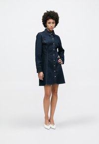 Τζιν φόρεμα σε σκούρο μπλε χρώμα με κουμπιά στο μπροστινό μέρος, γιακά και μακριά μανίκια. Διαθέτει αντίθετες ραφές και δομημένο σχήμα Α-γραμμή.