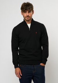 Schwarzer Reißverschluss-Pullover aus strukturiertem Stoff, mit hohem Kragen, gerippten Bündchen und einem kleinen roten Logo auf der Brust. Getragen mit blauen Jeans.