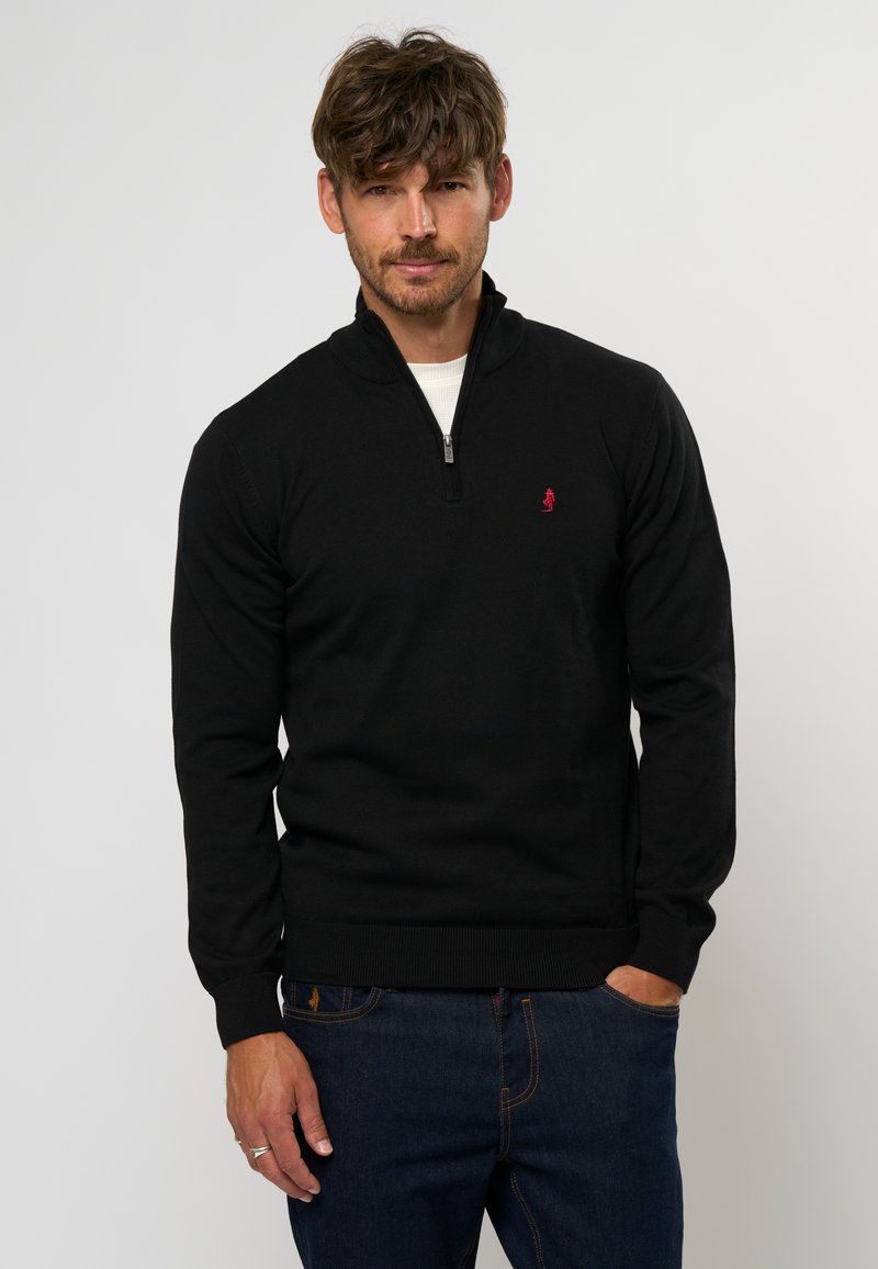 Zwarte zip-up trui van textuurstof, met een hoge kraag, ribbelmanchetten en een klein rood logo op de borst. Draagt het met blauwe spijkerbroek.