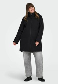 Chaqueta negra impermeable con cremallera frontal, capucha y bolsillos laterales. Presenta una cintura ligeramente ajustada y mangas largas. Combinada con jeans grises.