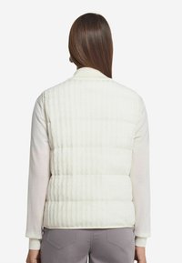 Gepolsterte Weste in Cremeweiß mit gerippter Textur. Das Design umfasst einen hohen Kragen und wird mit einem hellen Langarmtop kombiniert.