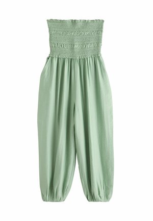 Kvinde med langt mørkt hår iført en lysegrøn, stropløs jumpsuit og sorte sandaler står vendt mod en hvid søjle og en træskodde dør.